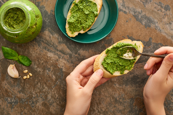 cropped view of woman adding pesto sauce with spoon on baguette slice on stone surface - Foto, Bild