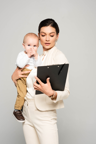 young businesswoman with baby boy on hands working with clipboard, isolated on grey - Foto, afbeelding