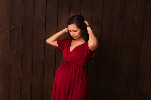 Kırmızı elbiseli hamile kadın koyu kahverengi arka planda el ele tutuşuyor. Hamilelik, annelik, beklenti konsepti. Hamileliğin güzel ve hassas fotoğrafı. - Fotoğraf, Görsel
