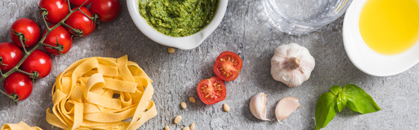top view of raw Pappardelle near tomatoes, garlic, basil, pine nuts, olive oil, water and pesto sauce on grey surface, panoramic shot - Valokuva, kuva