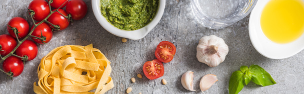 top view of raw Pappardelle near tomatoes, garlic, basil, pine nuts, olive oil, water and pesto sauce on grey surface, panoramic shot - Photo, Image