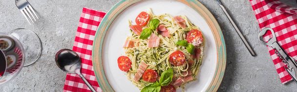 top view of Pappardelle with tomatoes, basil and prosciutto on plate on plaid napkin with cutlery near red wine on grey surface, panoramic shot - Фото, зображення