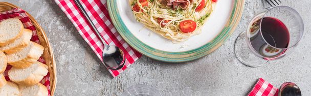 top view of served Pappardelle with tomatoes, basil and prosciutto near baguette, red wine on grey surface, panoramic shot - Photo, Image