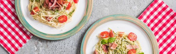 top view of served Pappardelle with tomatoes, basil and prosciutto on plaid napkins on grey surface, panoramic shot - 写真・画像