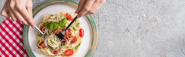 partial view of woman eating Pappardelle with tomatoes, basil and prosciutto on plaid napkin on grey surface, panoramic shot - Photo, Image