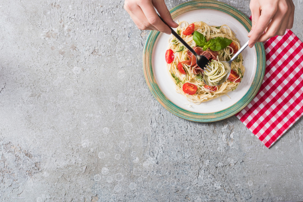 partial view of woman eating Pappardelle with tomatoes, basil and prosciutto on plaid napkin on grey surface - 写真・画像