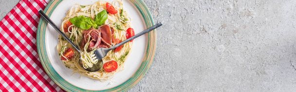 top view of served Pappardelle with tomatoes, basil and prosciutto on plaid napkin with cutlery on grey surface, panoramic shot - Фото, изображение