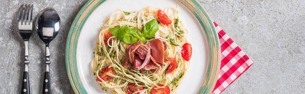 top view of served Pappardelle with tomatoes, basil and prosciutto on plaid napkin with cutlery on grey surface, panoramic shot - Foto, Bild