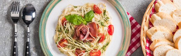 top view of served Pappardelle with tomatoes, basil and prosciutto near baguette and cutlery on grey surface, panoramic shot - Fotoğraf, Görsel