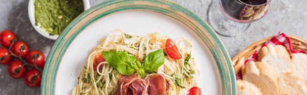 selective focus of served Pappardelle with tomatoes, basil and prosciutto near baguette, red wine, pesto, tomatoes on grey surface, panoramic shot - Фото, изображение