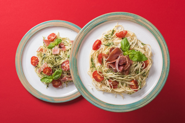 bovenaanzicht van gekookte Pappardelle met tomaten, basilicum en prosciutto op borden op rode achtergrond - Foto, afbeelding