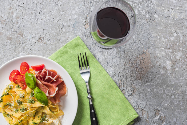 top view of cooked Pappardelle with tomatoes, basil and prosciutto near red wine on grey surface - 写真・画像