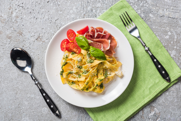 top view of cooked Pappardelle with tomatoes, basil and prosciutto near cutlery on grey surface - Fotoğraf, Görsel