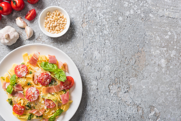 Sarımsak, çam fıstığı ve gri yüzeyde domatesli fesleğen ve prosciutto ile pişmiş Pappardelle 'in üst görüntüsü - Fotoğraf, Görsel