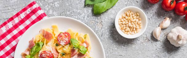 top view of cooked Pappardelle with tomatoes, basil and prosciutto on plaid napkin near ingredients on grey surface, panoramic shot - Photo, Image