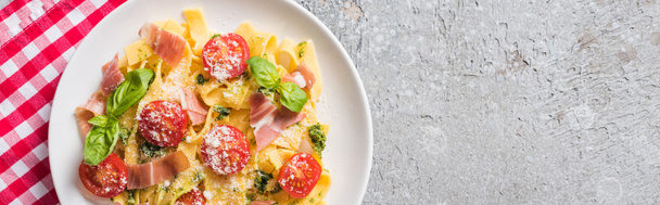 top view of cooked Pappardelle with tomatoes, basil and prosciutto on plaid napkin on grey surface, panoramic shot - Photo, Image