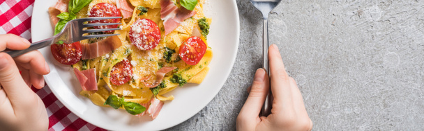 partial view of woman eating Pappardelle with tomatoes, basil and prosciutto on plaid napkin on grey surface, panoramic shot - Foto, imagen