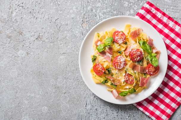top view of cooked Pappardelle with tomatoes, basil and prosciutto on plaid napkin on grey surface - Photo, Image