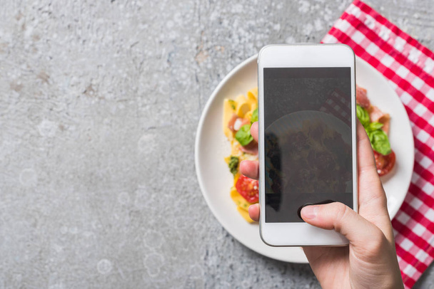 vista parziale della donna che fotografa Pappardelle con pomodori, basilico e prosciutto su smartphone su superficie grigia
 - Foto, immagini