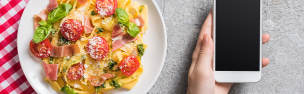 partial view of woman holding smartphone near Pappardelle with tomatoes, basil and prosciutto on plaid napkin on grey surface, panoramic shot - Photo, Image