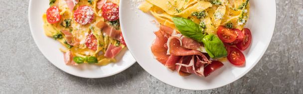 selective focus of cooked Pappardelle with tomatoes, basil and prosciutto on plates on grey surface, panoramic shot - Photo, Image