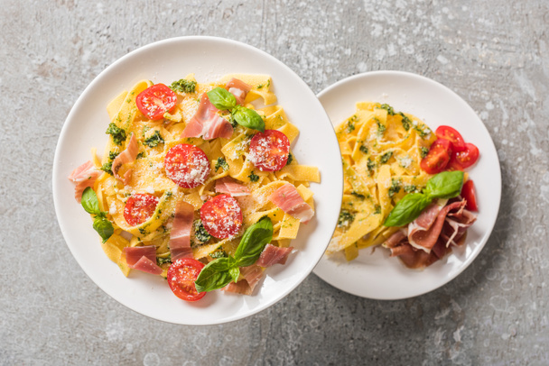 selective focus of cooked Pappardelle with tomatoes, basil and prosciutto on plates on grey surface - 写真・画像