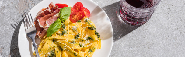 Draufsicht auf schmackhafte Pappardelle mit Tomaten, Pesto und Prosciutto mit Gabel in Rotweinnähe auf grauer Oberfläche, Panoramaaufnahme - Foto, Bild