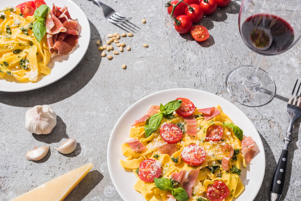 fresh Pappardelle with tomatoes, pesto and prosciutto near red wine and ingredients on grey surface - 写真・画像