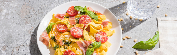 tasty Pappardelle with tomatoes, pesto and prosciutto near water, napkin and pine nuts on grey surface, panoramic shot - Foto, imagen