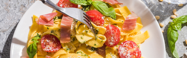 top view of tasty Pappardelle with tomatoes, pesto and prosciutto with fork on grey surface, panoramic shot - Fotó, kép