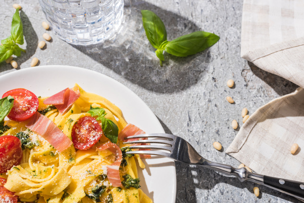 top view of tasty Pappardelle with tomatoes, pesto and prosciutto with fork near water and pine nuts on grey surface - Foto, immagini
