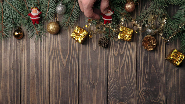 Ano Novo e decoração de Natal. Mão coloca alguns presentes, bolas de Natal, cones de pinho e pequenos brinquedos de Papai Noel perto dos galhos da árvore de Natal em uma mesa de madeira. Vista superior
 - Filmagem, Vídeo