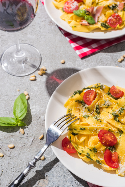 tasty Pappardelle with tomatoes, pesto and prosciutto with fork near red wine, basil and pine nuts on grey surface - Photo, Image