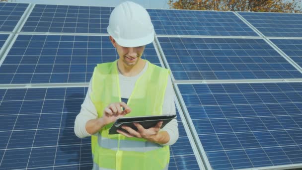 A worker in a helmet works with a tablet near a ground-based solar power station - Felvétel, videó