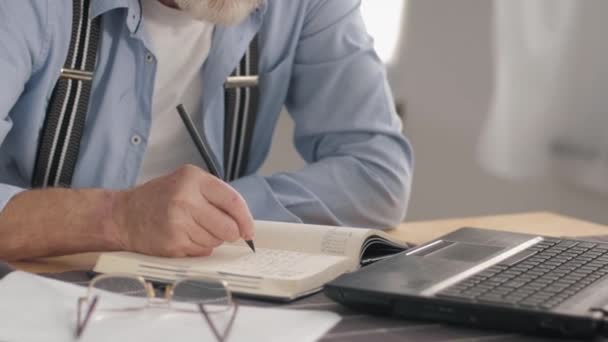 Porträt eines modernen Rentners, der zu Hause am tragbaren Computer arbeitet und Rechnungen in sein Notizbuch schreibt, lächelnd in die Kamera blickt - Filmmaterial, Video