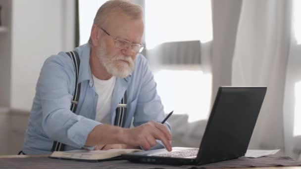 πορτραίτο ενός ηλικιωμένου άνδρα με γυαλιά με γενειάδα που εργάζεται στον υπολογιστή και αναλύει τραπεζικούς λογαριασμούς για να πληρώσει online κάθεται στο φορητό υπολογιστή στο τραπέζι - Πλάνα, βίντεο