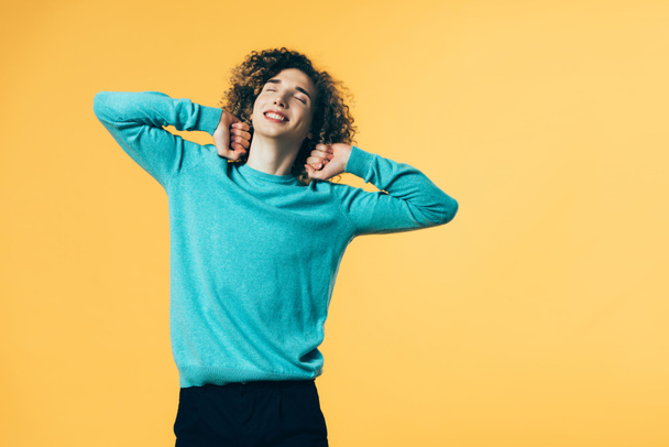 smiling curly teenager stretching himself with closed eyes isolated on yellow - Foto, immagini