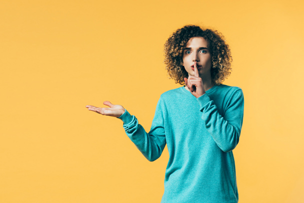curly teenager showing shh gesture and pointing with hand aside isolated on yellow - Foto, Imagen