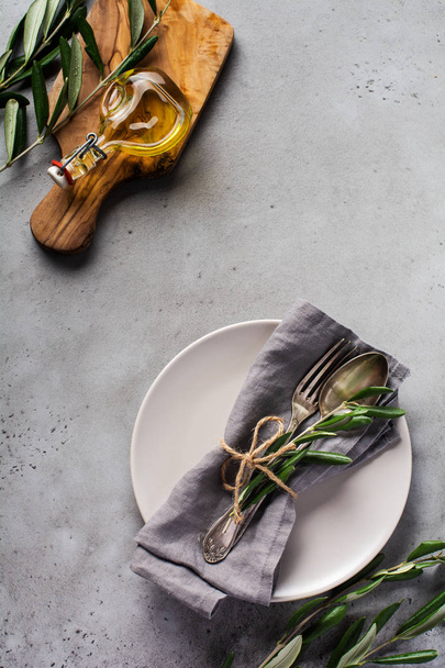 Rustic vintage set of cutlery. Plate with grey linen napkin, fork and spoon, olive tree branch over rustic concrete gray old background. Fall holiday table decoration setting. Top view, copy space. - Photo, Image