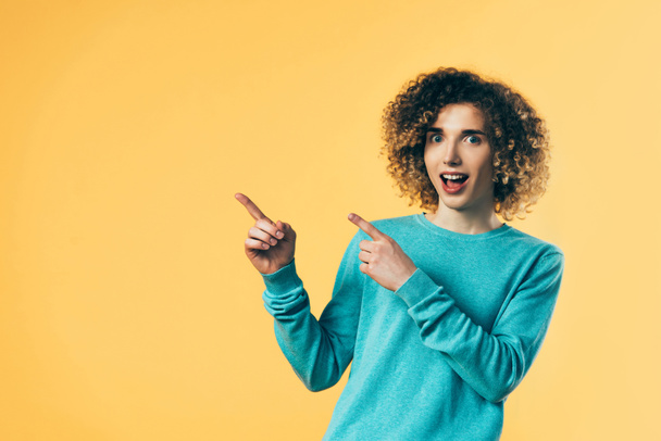 curly teenager with open mouth pointing with fingers aside isolated on yellow - Photo, Image
