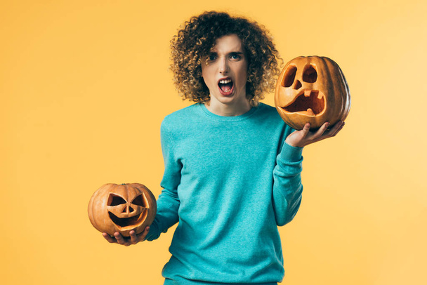 scared curly teenager holding Halloween pumpkins and screaming isolated on yellow - Photo, Image