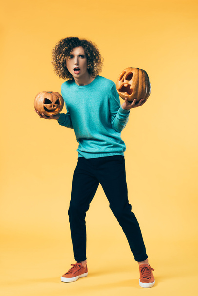 scared curly teenager holding Halloween pumpkins on yellow - Foto, imagen