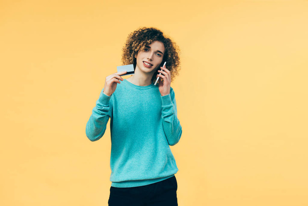 кучерявий підліток говорить на смартфоні і тримає кредитну картку ізольовано на жовтому
 - Фото, зображення