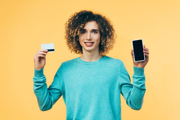 smiling curly teenager holding smartphone and credit card isolated on yellow - 写真・画像