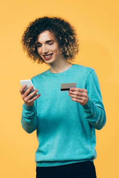 smiling curly teenager holding smartphone and credit card isolated on yellow - Foto, immagini