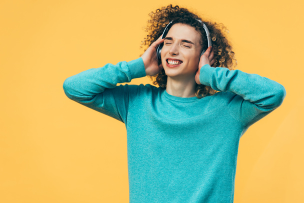smiling curly teenager listening music in headphones with closed eyes isolated on yellow - Фото, зображення