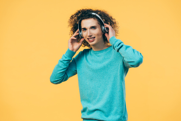 smiling curly teenager listening music in headphones isolated on yellow - Photo, Image