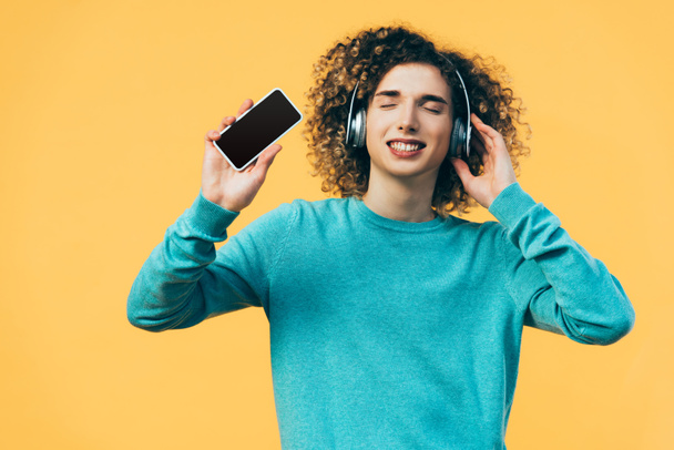 curly teenager with closed eyes holding smartphone and listening music in headphones isolated on yellow - Foto, afbeelding