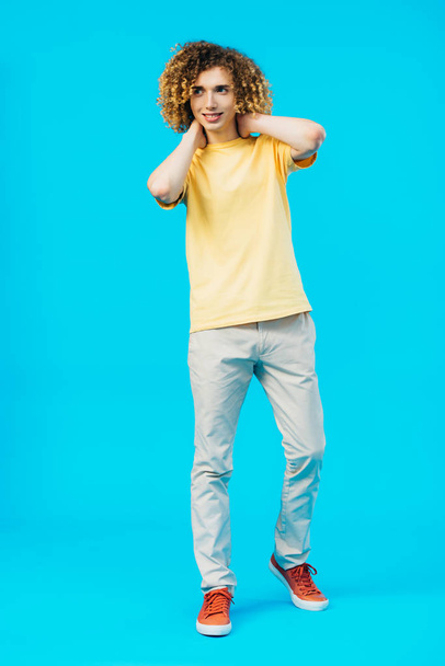 smiling curly teenager posing with hands behind neck isolated on blue - Foto, Imagem