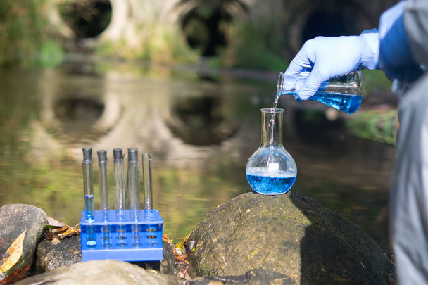 wetenschapper hand, in een beschermend pak en handschoenen, giet blauwe vloeistof in een kolf, tegen de achtergrond van rioolbuizen - Foto, afbeelding